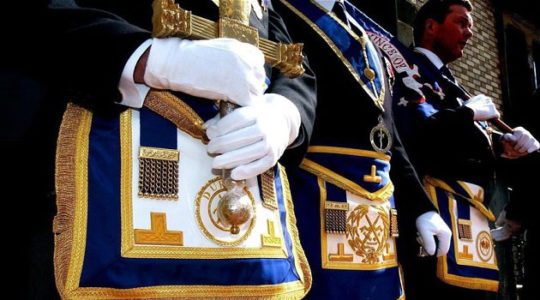 Masones vestidos con guantes blancos, mandiles ceremoniales y espadas durante un acto público.