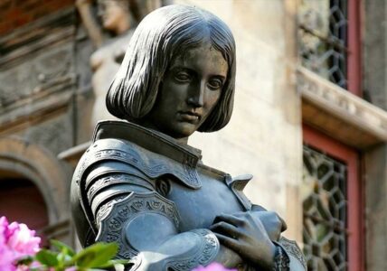 Estatua de Juana de Arco frente a una iglesia, símbolo de su conflicto con la jerarquía eclesiástica