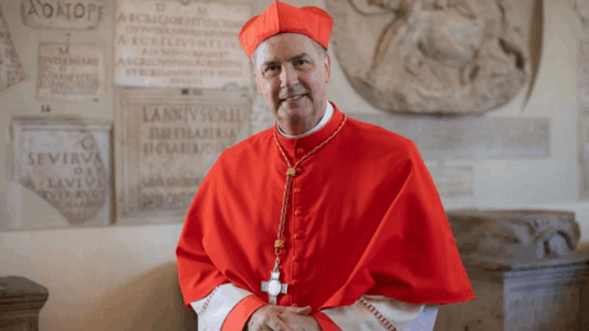 El cardenal español Ángel Artime vestido con traje rojo cardenalicio frente a un muro con inscripciones religiosas en Asís.